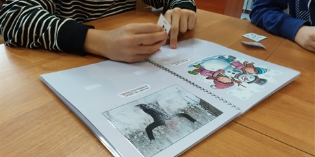 Powiększ grafikę: Na stole leży otwarta książka w formie skoroszytu, w niej obrazki bałwana i osoby na śniegu. Widać ręce.