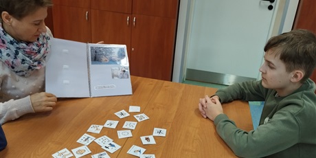 Powiększ grafikę: Na stole leżą symbole PCS, kobieta pokazuje otwary skoroszyt, dwóch chłopców siedzi przy stole.