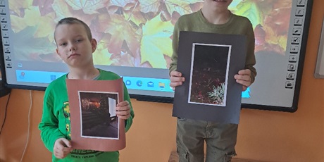 Powiększ grafikę: Dwóch uczniów stoi i trzyma w rękach swoje konkursowe prace. Jeden z uczniów stoi na podeście. Na drugim planie tło złożone z jesiennych liści. 