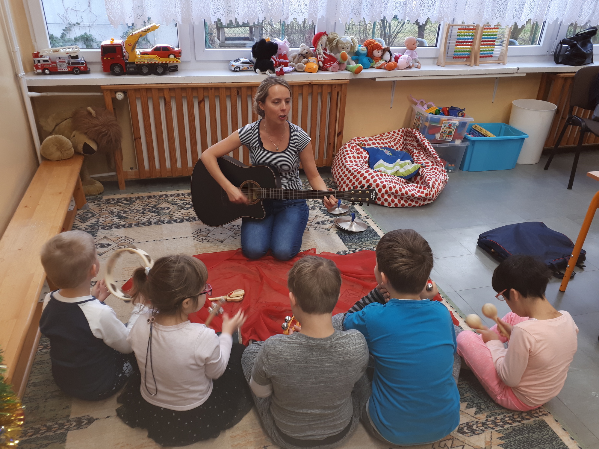 Kobieta z gitarą siedząca na dywanie a przed nią piątka dzieci z różnymi instrumentami muzycznymi.