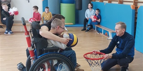 Powiększ grafikę: Chłopiec na wózku z pomocą nauczycielki próbuje wrzucić piłkę do kosza, który trzyma przed nim nauczyciel. Dalej na ławeczkach siedzą inni uczniowie.