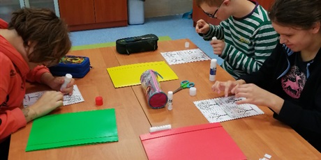 Powiększ grafikę: Dwie dziewczynki i chłopiec siedzą przy stole, na którym leżą kolorowe teczki i kartki. Przyklejają na kartki elementy.