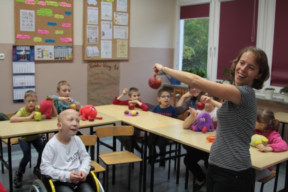 Dzieci siedzące w sali lekcyjnej i kobieta trzymająca jabłko
